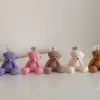 It looks like the image shows five teddy bear–shaped candles in different colors (purple, pink, light brown, cream, and medium brown) sitting in a row on a white surface. Each candle has a wick on top with a small tag that says 