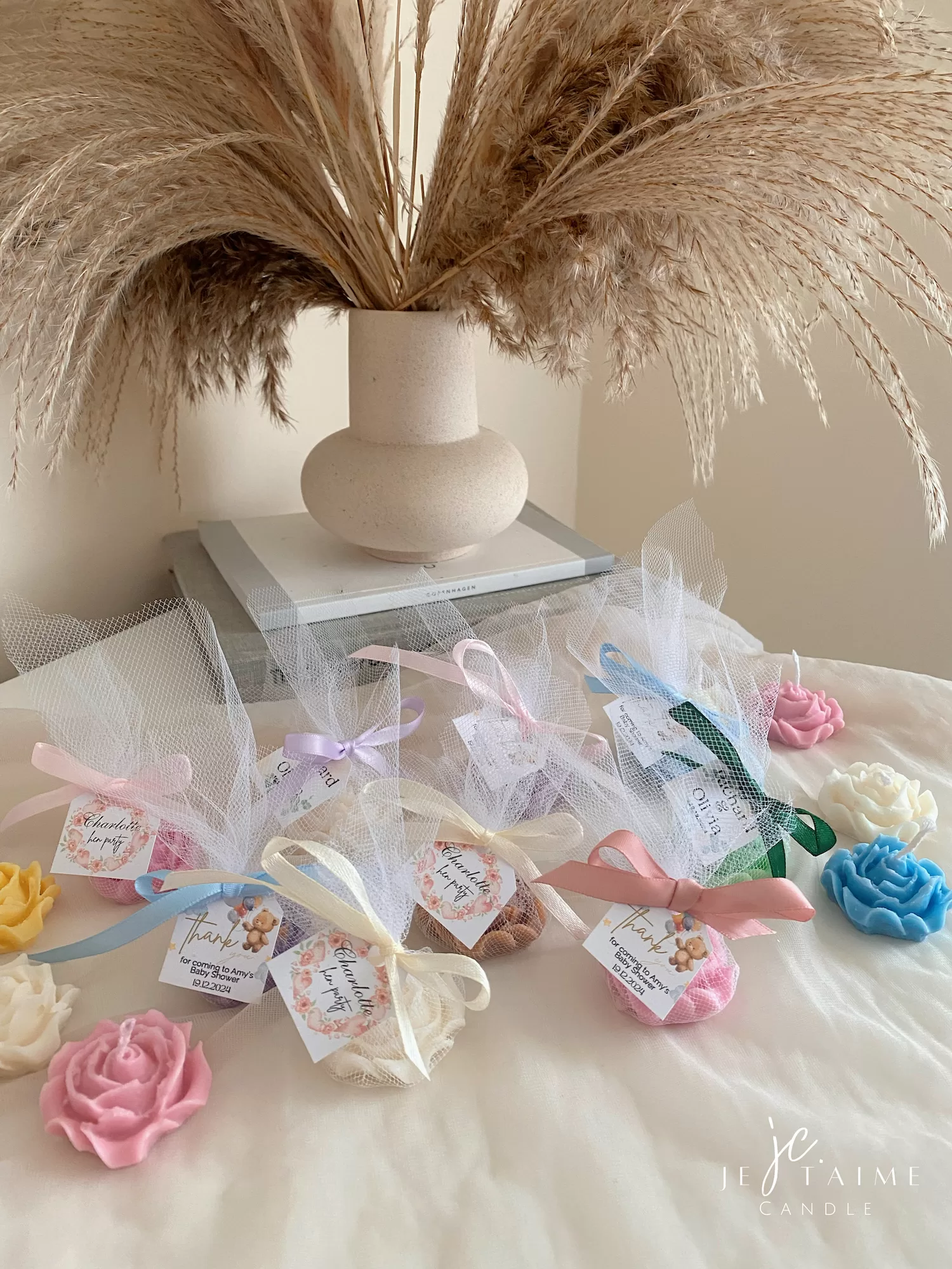 Colorful rose candles in pink, blue, yellow, and white, wrapped in white organza bags with personalized 'Thank You' tags for Amy's baby shower on 12/01/2024, displayed with pampas grass decor in the background