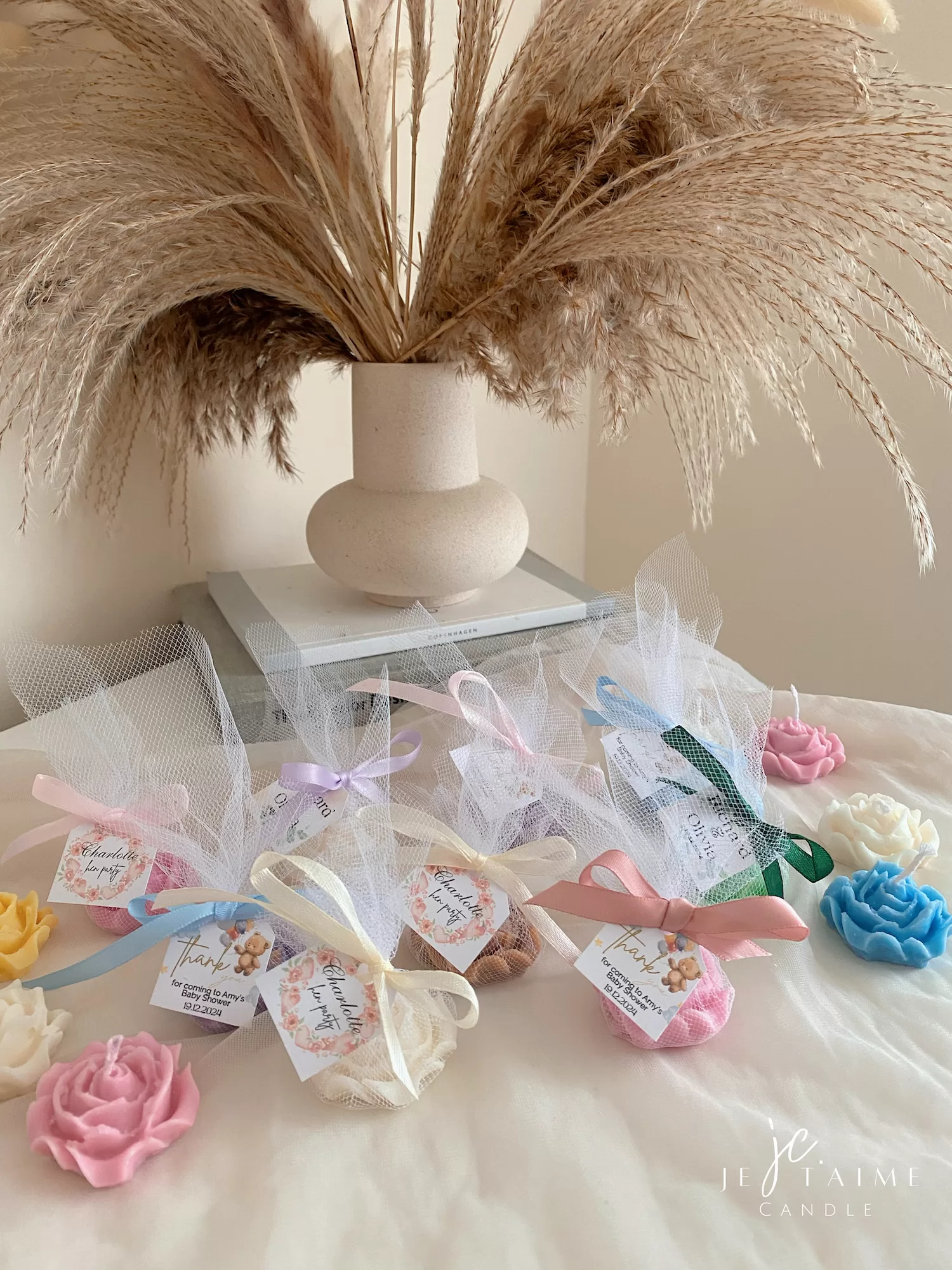 Colorful rose candles in pink, blue, yellow, and white, wrapped in white organza bags with personalized 'Thank You' tags for Amy's baby shower on 12/01/2024, displayed with pampas grass decor in the background