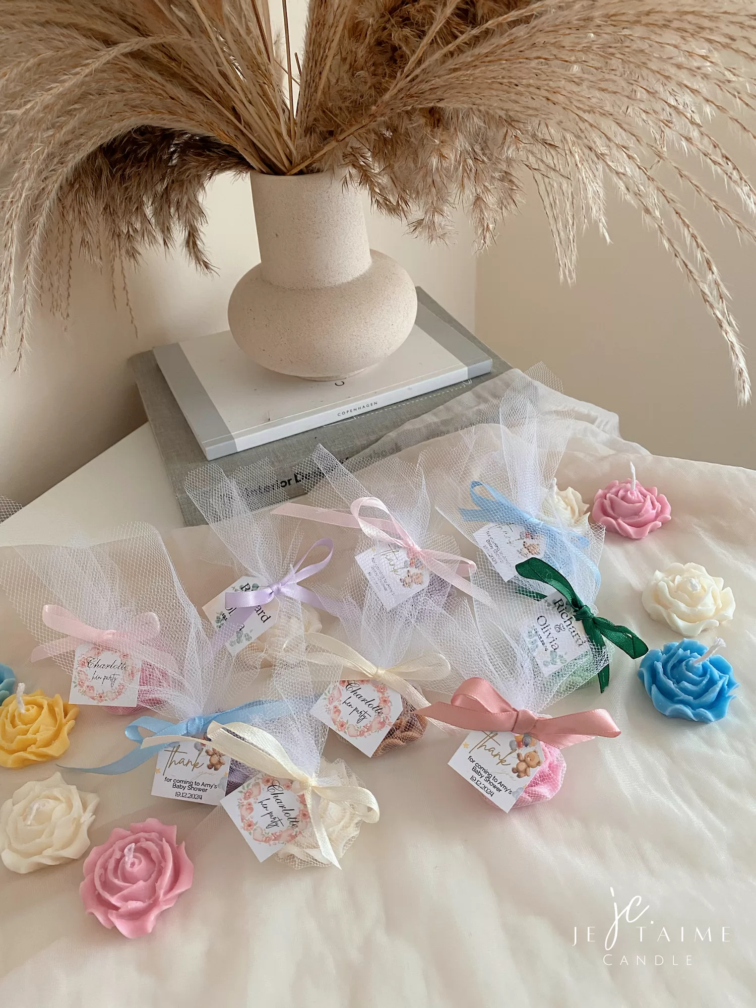 Colorful rose candles in pink, blue, yellow, and white, wrapped in white organza bags with personalized 'Thank You' tags for Amy's baby shower on 12/01/2024, displayed with pampas grass decor in the background
