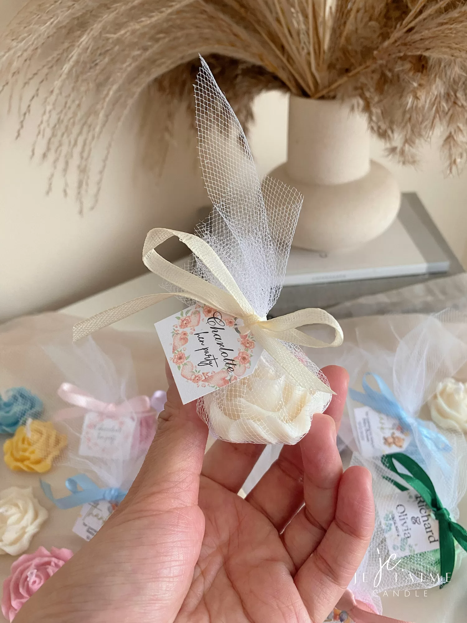 Colorful rose candles in pink, blue, yellow, and white, wrapped in white organza bags with personalized 'Thank You' tags for Amy's baby shower on 12/01/2024, displayed with pampas grass decor in the background