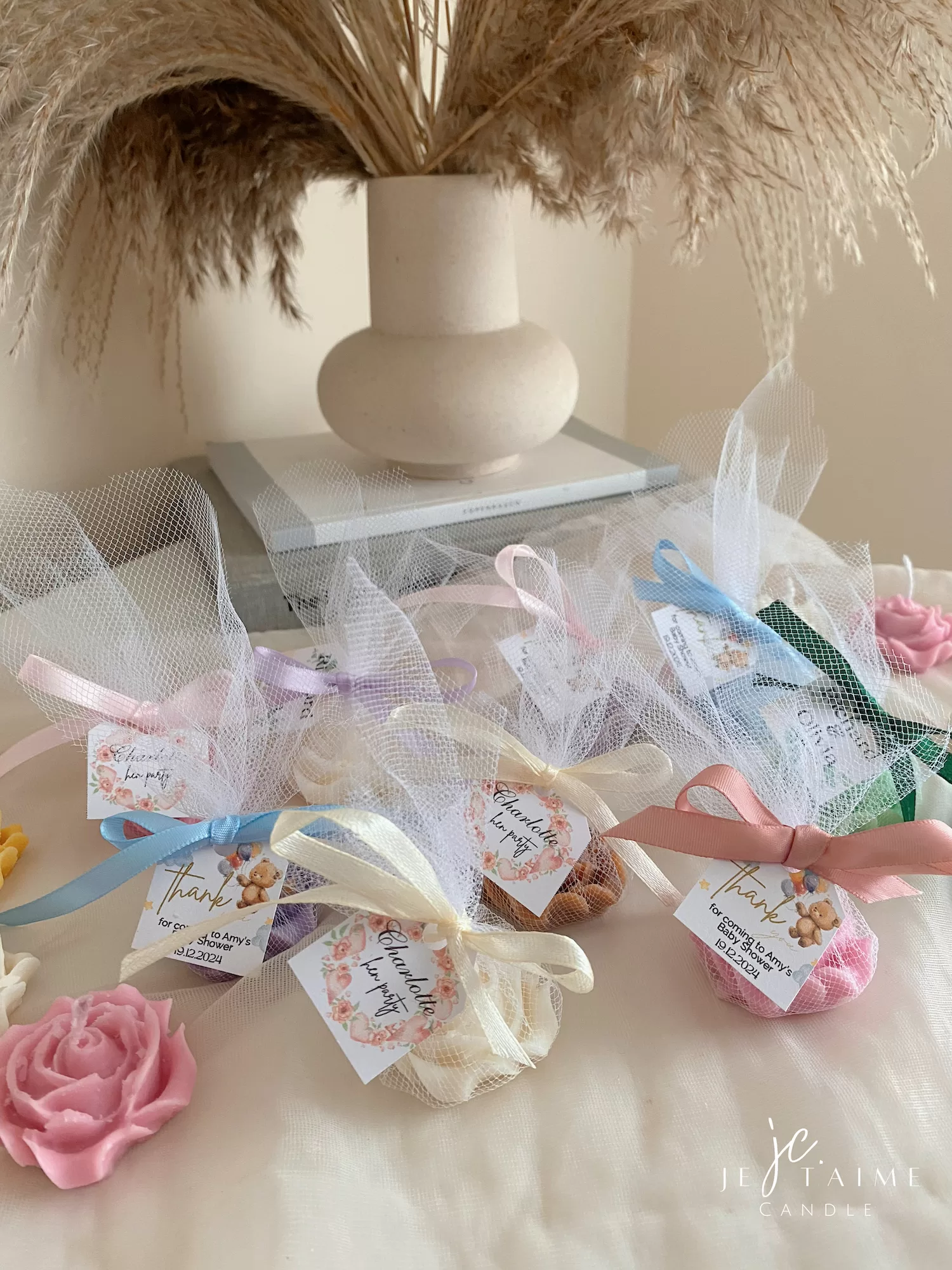 Colorful rose candles in pink, blue, yellow, and white, wrapped in white organza bags with personalized 'Thank You' tags for Amy's baby shower on 12/01/2024, displayed with pampas grass decor in the background