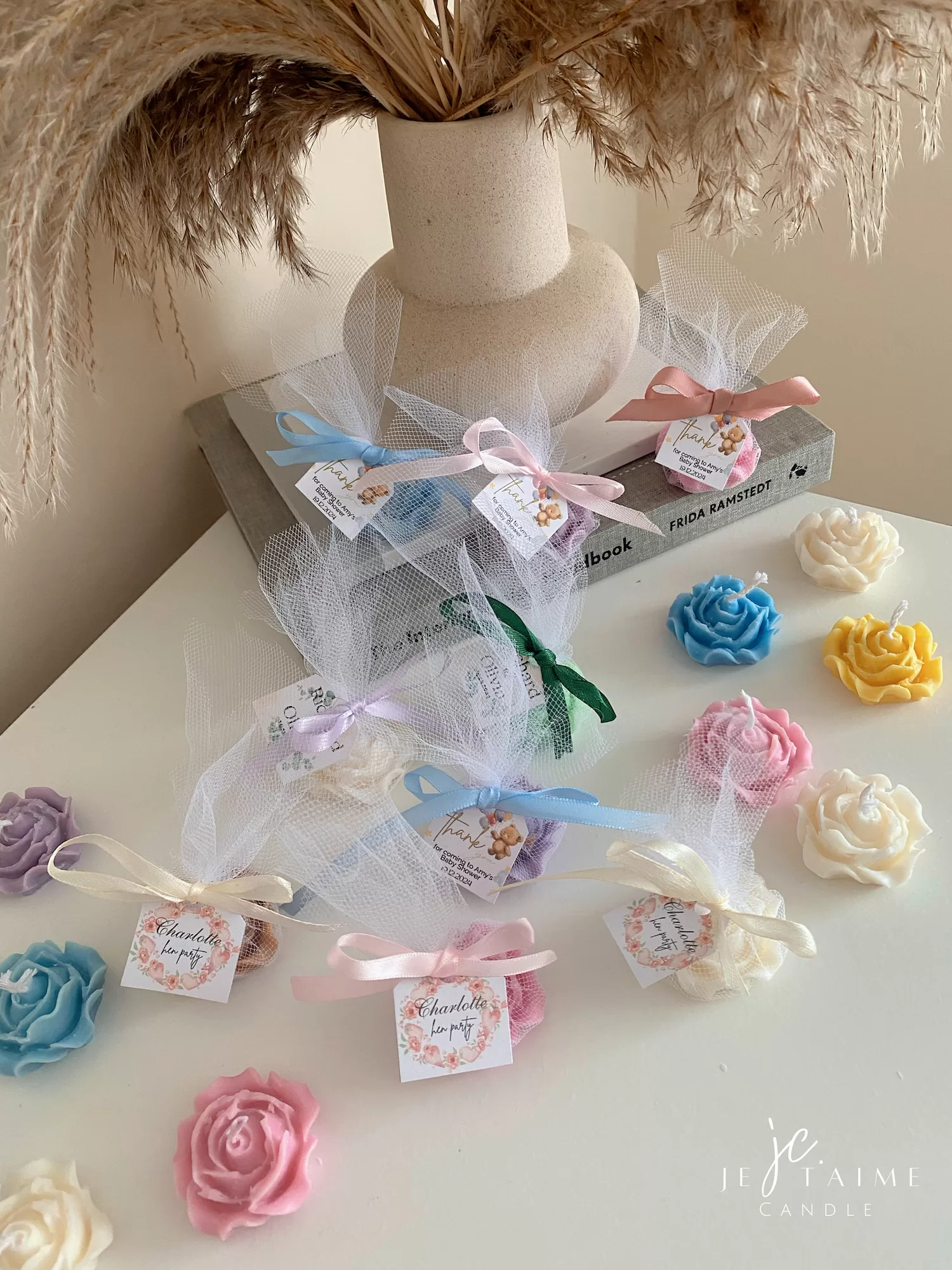 Colorful rose candles in pink, blue, yellow, and white, wrapped in white organza bags with personalized 'Thank You' tags for Amy's baby shower on 12/01/2024, displayed with pampas grass decor in the background
