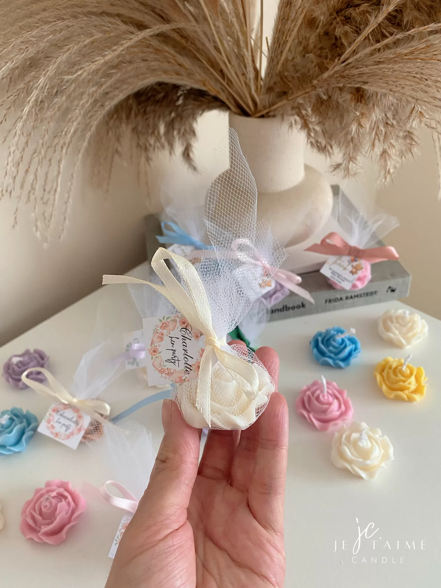 Colorful rose candles in pink, blue, yellow, and white, wrapped in white organza bags with personalized 'Thank You' tags for Amy's baby shower on 12/01/2024, displayed with pampas grass decor in the background