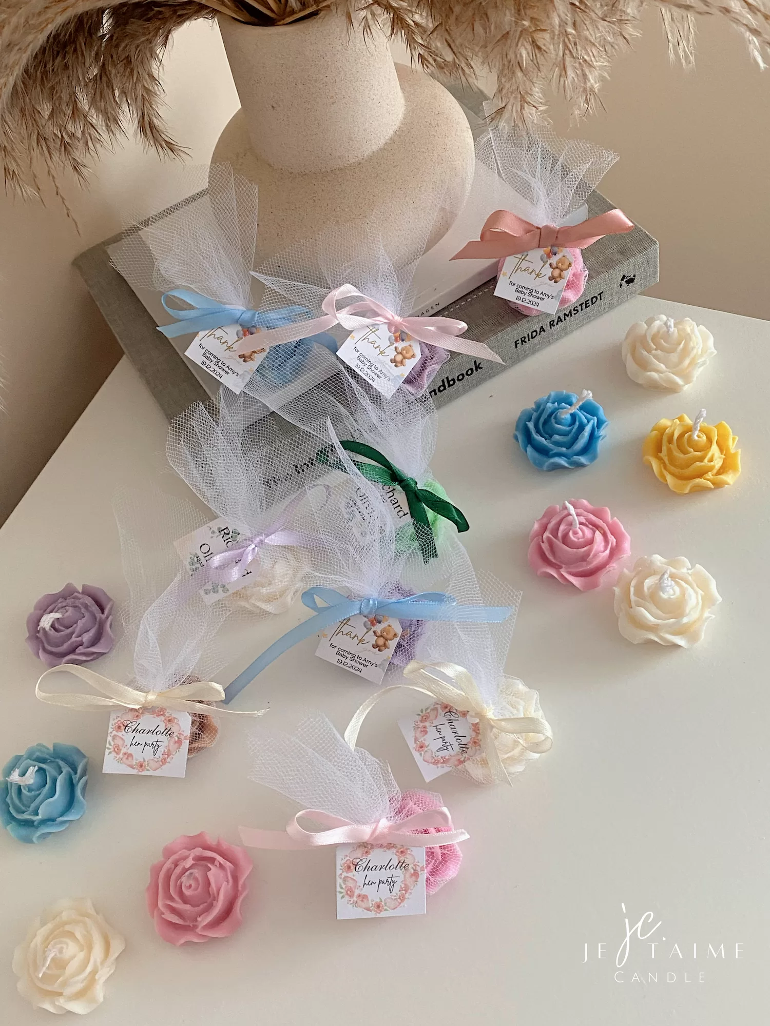 Colorful rose candles in pink, blue, yellow, and white, wrapped in white organza bags with personalized 'Thank You' tags for Amy's baby shower on 12/01/2024, displayed with pampas grass decor in the background