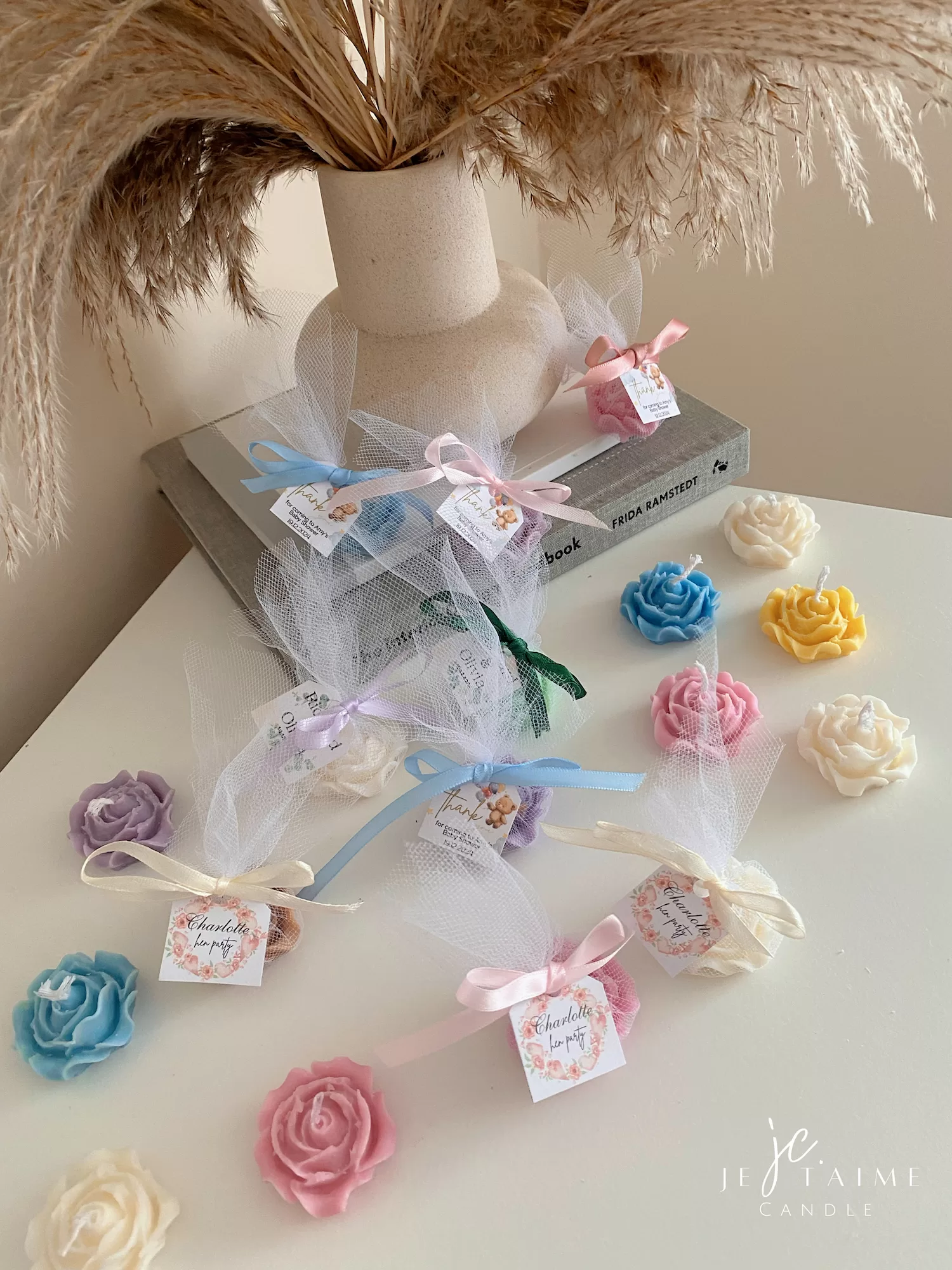 Colorful rose candles in pink, blue, yellow, and white, wrapped in white organza bags with personalized 'Thank You' tags for Amy's baby shower on 12/01/2024, displayed with pampas grass decor in the background