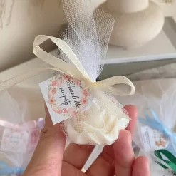 A small white rose-shaped candle wrapped in white tulle and tied with a cream ribbon, with a floral wreath tag reading ‘Charlotte’s hen party’ held in a person’s hand.