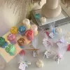 This image shows an assortment of small, flower-shaped candles that resemble peony buds in various colors—yellow, blue, white, pink, green, and brown. Some are displayed on a clear iridescent stand, while others are individually wrapped in white tulle and tied with pastel-colored ribbons (blue, pink, cream, brown). Each wrapped candle has a small personalized tag with messages like “Thank you” or wedding names. In the background, there’s a decorative vase with pampas grass placed on top of stacked books. The branding “JE T’AIME CANDLE” is visible in the bottom right corner.