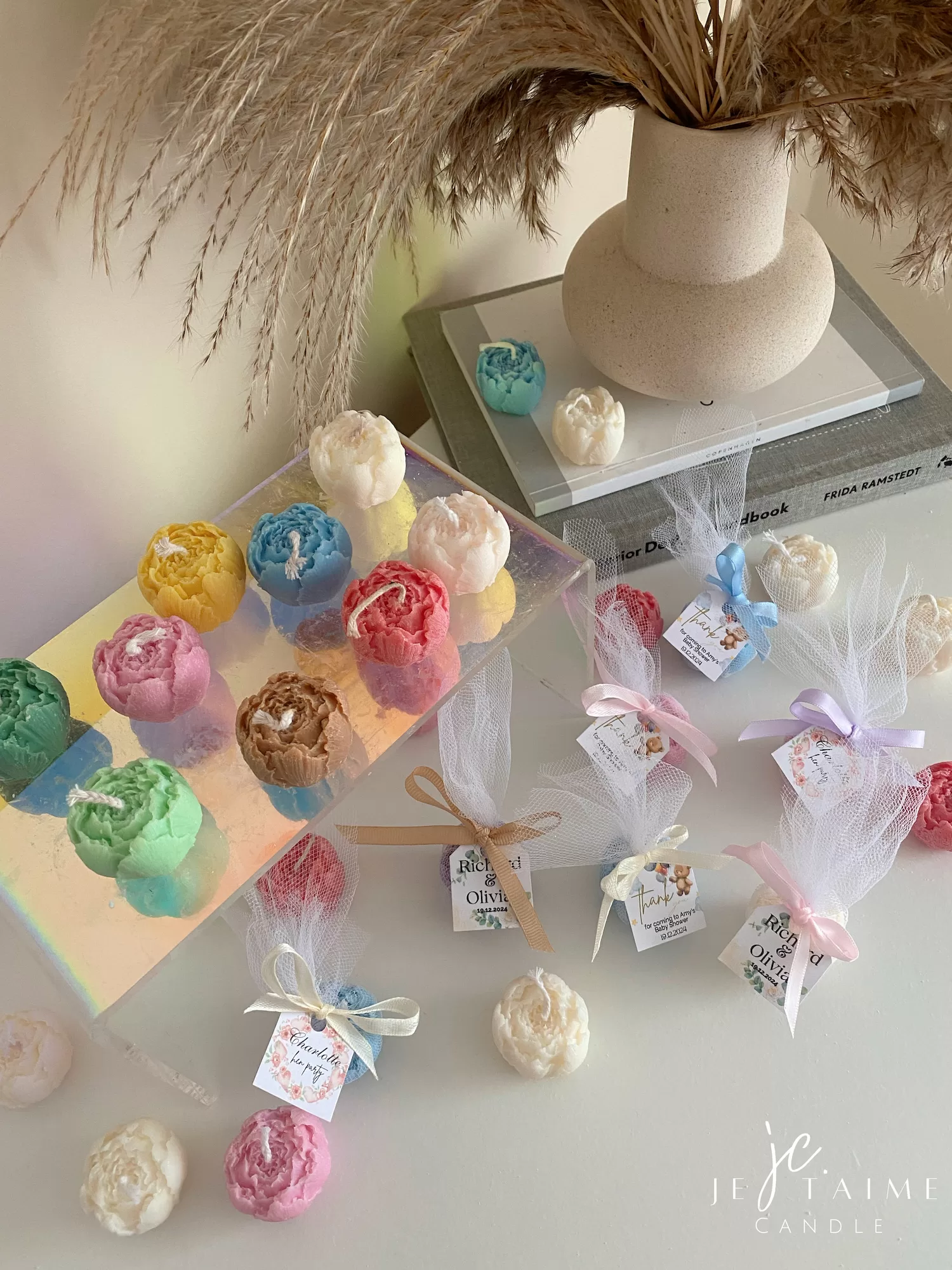 This image shows an assortment of small, flower-shaped candles that resemble peony buds in various colors—yellow, blue, white, pink, green, and brown. Some are displayed on a clear iridescent stand, while others are individually wrapped in white tulle and tied with pastel-colored ribbons (blue, pink, cream, brown). Each wrapped candle has a small personalized tag with messages like “Thank you” or wedding names. In the background, there’s a decorative vase with pampas grass placed on top of stacked books. The branding “JE T’AIME CANDLE” is visible in the bottom right corner.