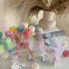 This image shows an assortment of small, flower-shaped candles that resemble peony buds in various colors—yellow, blue, white, pink, green, and brown. Some are displayed on a clear iridescent stand, while others are individually wrapped in white tulle and tied with pastel-colored ribbons (blue, pink, cream, brown). Each wrapped candle has a small personalized tag with messages like “Thank you” or wedding names. In the background, there’s a decorative vase with pampas grass placed on top of stacked books. The branding “JE T’AIME CANDLE” is visible in the bottom right corner.