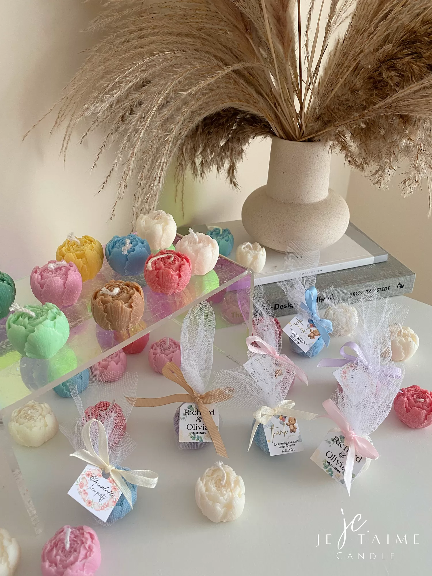 This image shows an assortment of small, flower-shaped candles that resemble peony buds in various colors—yellow, blue, white, pink, green, and brown. Some are displayed on a clear iridescent stand, while others are individually wrapped in white tulle and tied with pastel-colored ribbons (blue, pink, cream, brown). Each wrapped candle has a small personalized tag with messages like “Thank you” or wedding names. In the background, there’s a decorative vase with pampas grass placed on top of stacked books. The branding “JE T’AIME CANDLE” is visible in the bottom right corner.