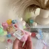 This image shows an assortment of small, flower-shaped candles that resemble peony buds in various colors—yellow, blue, white, pink, green, and brown. Some are displayed on a clear iridescent stand, while others are individually wrapped in white tulle and tied with pastel-colored ribbons (blue, pink, cream, brown). Each wrapped candle has a small personalized tag with messages like “Thank you” or wedding names. In the background, there’s a decorative vase with pampas grass placed on top of stacked books. The branding “JE T’AIME CANDLE” is visible in the bottom right corner.