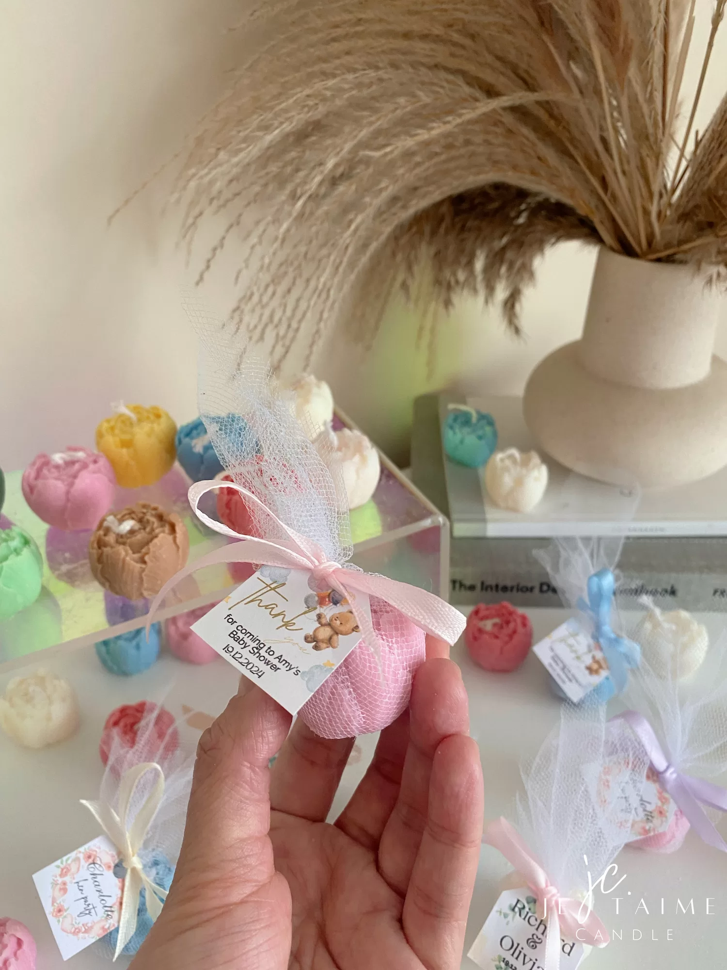 This image shows an assortment of small, flower-shaped candles that resemble peony buds in various colors—yellow, blue, white, pink, green, and brown. Some are displayed on a clear iridescent stand, while others are individually wrapped in white tulle and tied with pastel-colored ribbons (blue, pink, cream, brown). Each wrapped candle has a small personalized tag with messages like “Thank you” or wedding names. In the background, there’s a decorative vase with pampas grass placed on top of stacked books. The branding “JE T’AIME CANDLE” is visible in the bottom right corner.