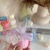 This image shows an assortment of small, flower-shaped candles that resemble peony buds in various colors—yellow, blue, white, pink, green, and brown. Some are displayed on a clear iridescent stand, while others are individually wrapped in white tulle and tied with pastel-colored ribbons (blue, pink, cream, brown). Each wrapped candle has a small personalized tag with messages like “Thank you” or wedding names. In the background, there’s a decorative vase with pampas grass placed on top of stacked books. The branding “JE T’AIME CANDLE” is visible in the bottom right corner.