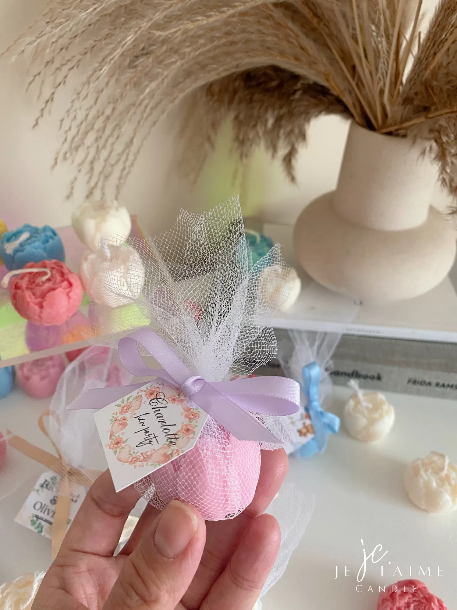 This image shows an assortment of small, flower-shaped candles that resemble peony buds in various colors—yellow, blue, white, pink, green, and brown. Some are displayed on a clear iridescent stand, while others are individually wrapped in white tulle and tied with pastel-colored ribbons (blue, pink, cream, brown). Each wrapped candle has a small personalized tag with messages like “Thank you” or wedding names. In the background, there’s a decorative vase with pampas grass placed on top of stacked books. The branding “JE T’AIME CANDLE” is visible in the bottom right corner.