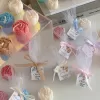 This image shows an assortment of small, flower-shaped candles that resemble peony buds in various colors—yellow, blue, white, pink, green, and brown. Some are displayed on a clear iridescent stand, while others are individually wrapped in white tulle and tied with pastel-colored ribbons (blue, pink, cream, brown). Each wrapped candle has a small personalized tag with messages like “Thank you” or wedding names. In the background, there’s a decorative vase with pampas grass placed on top of stacked books. The branding “JE T’AIME CANDLE” is visible in the bottom right corner.