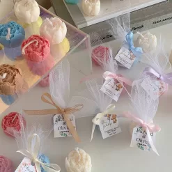 This image shows an assortment of small, flower-shaped candles that resemble peony buds in various colors—yellow, blue, white, pink, green, and brown. Some are displayed on a clear iridescent stand, while others are individually wrapped in white tulle and tied with pastel-colored ribbons (blue, pink, cream, brown). Each wrapped candle has a small personalized tag with messages like “Thank you” or wedding names. In the background, there’s a decorative vase with pampas grass placed on top of stacked books. The branding “JE T’AIME CANDLE” is visible in the bottom right corner.