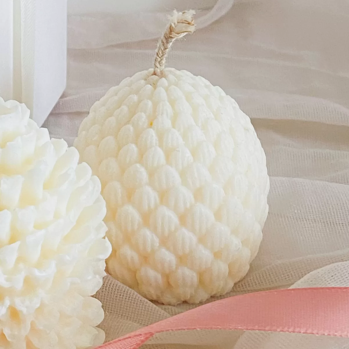 Pine cone-shaped candles displayed, set on soft fabric with a pink ribbon accent.