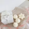 Three ivory pine cone-shaped and snowflake candles displayed beside a white gift box tied with a ribbon, set on soft fabric with a pink ribbon accent.