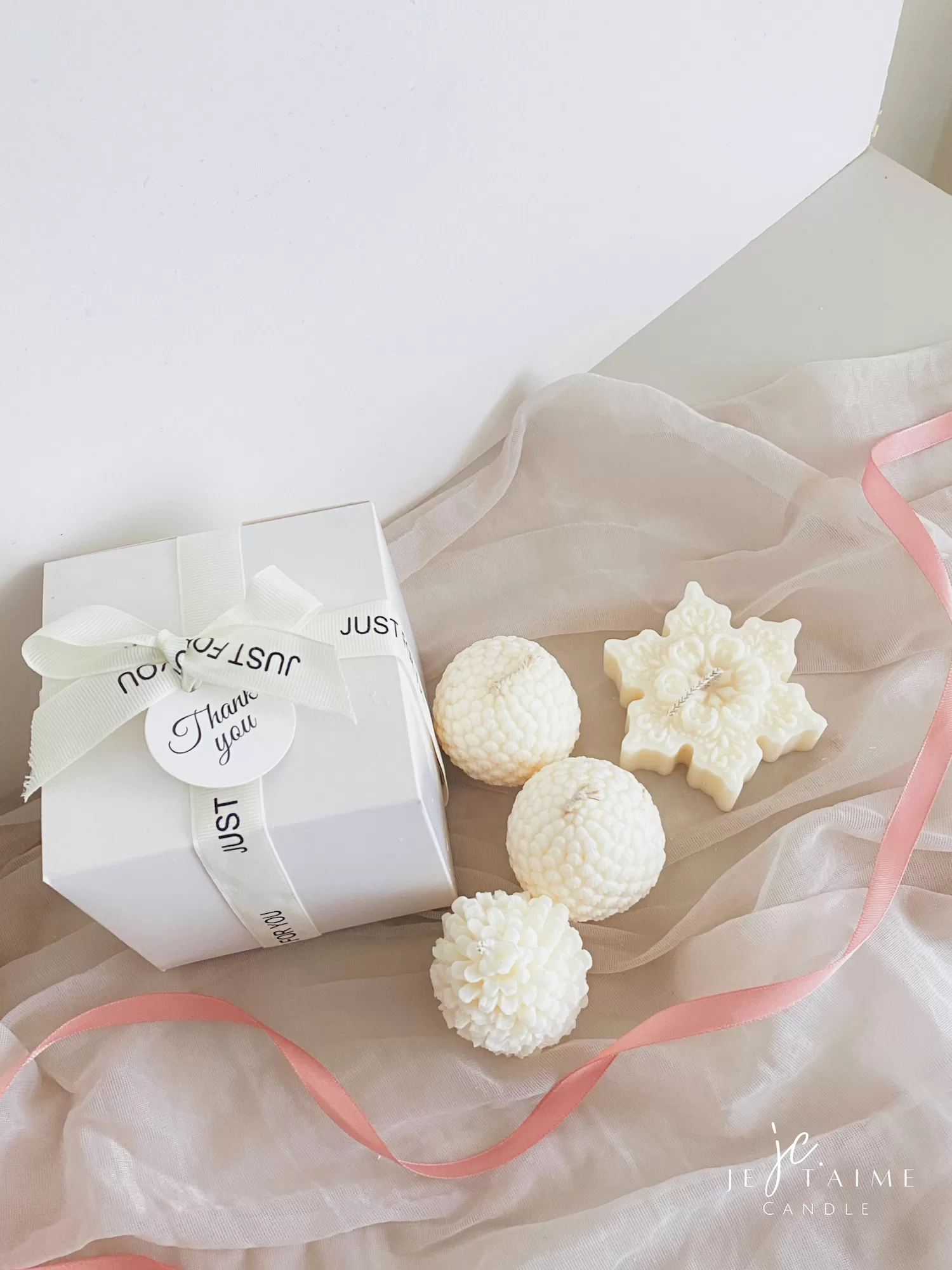 Three ivory pine cone-shaped and snowflake candles displayed beside a white gift box tied with a ribbon, set on soft fabric with a pink ribbon accent.