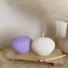 Two seashell-shaped candles in pastel tones, one lavender purple and one ivory white, displayed on a wooden tray with matches beside them. These handmade soy wax seashell candles feature a ribbed shell design, perfect as coastal home décor, beach wedding decorations, summer party favors, or unique handmade gifts. Minimalist and aesthetic, these shell candles bring a serene seaside vibe to any room.