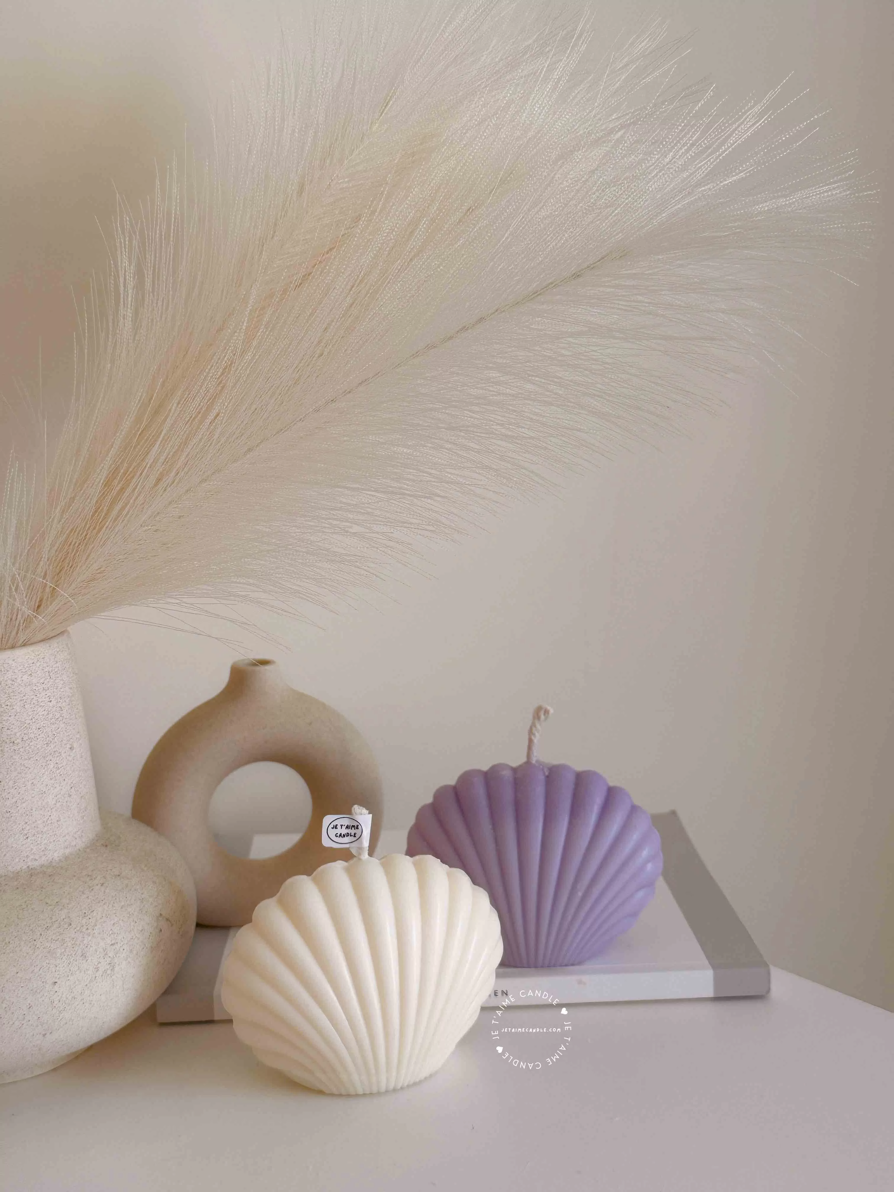 Two seashell-shaped candles in pastel tones, one lavender purple and one ivory white, displayed on a wooden tray with matches beside them. These handmade soy wax seashell candles feature a ribbed shell design, perfect as coastal home décor, beach wedding decorations, summer party favors, or unique handmade gifts. Minimalist and aesthetic, these shell candles bring a serene seaside vibe to any room.