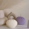Two seashell-shaped candles in pastel tones, one lavender purple and one ivory white, displayed on a wooden tray with matches beside them. These handmade soy wax seashell candles feature a ribbed shell design, perfect as coastal home décor, beach wedding decorations, summer party favors, or unique handmade gifts. Minimalist and aesthetic, these shell candles bring a serene seaside vibe to any room.