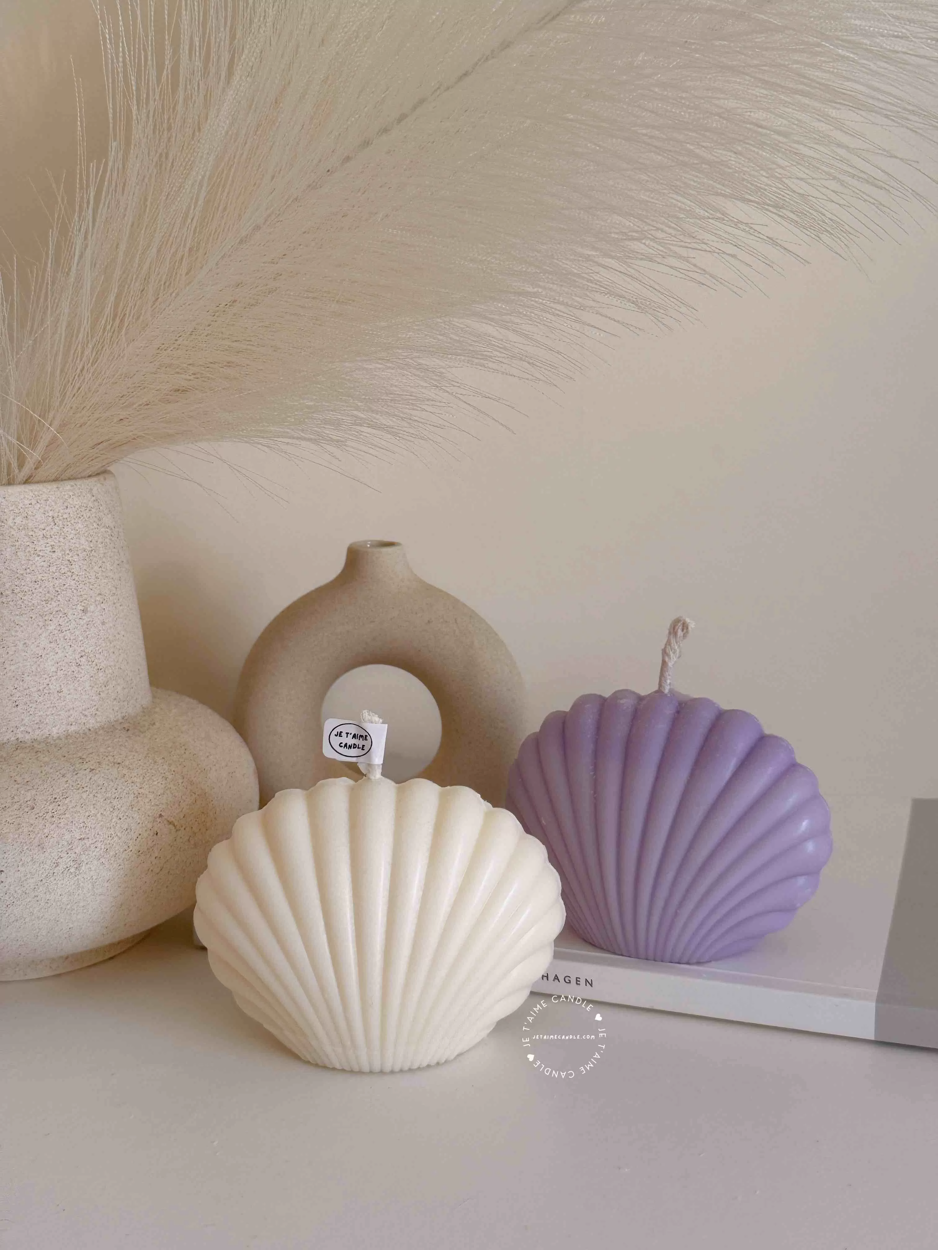 Two seashell-shaped candles in pastel tones, one lavender purple and one ivory white, displayed on a wooden tray with matches beside them. These handmade soy wax seashell candles feature a ribbed shell design, perfect as coastal home décor, beach wedding decorations, summer party favors, or unique handmade gifts. Minimalist and aesthetic, these shell candles bring a serene seaside vibe to any room.