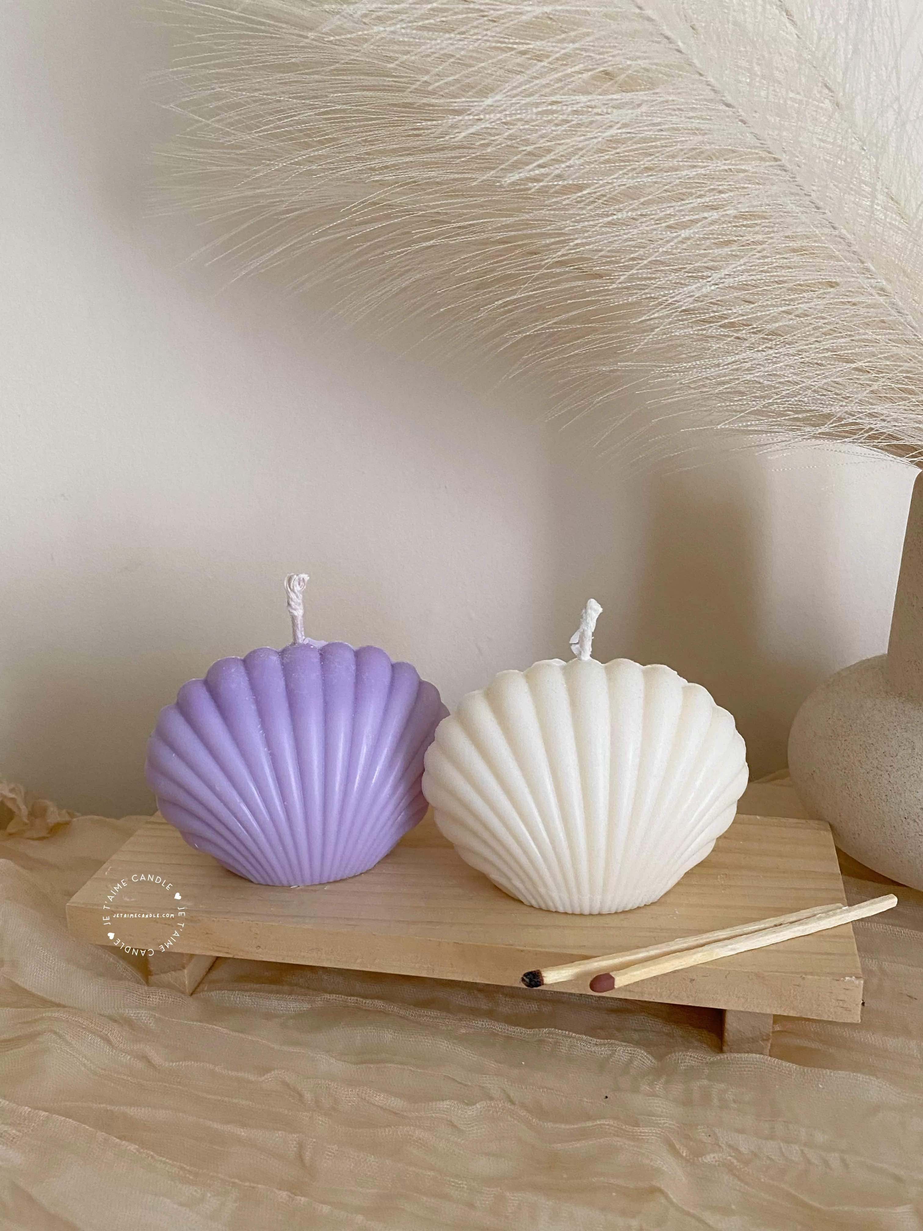 Two seashell-shaped candles in pastel tones, one lavender purple and one ivory white, displayed on a wooden tray with matches beside them. These handmade soy wax seashell candles feature a ribbed shell design, perfect as coastal home décor, beach wedding decorations, summer party favors, or unique handmade gifts. Minimalist and aesthetic, these shell candles bring a serene seaside vibe to any room.