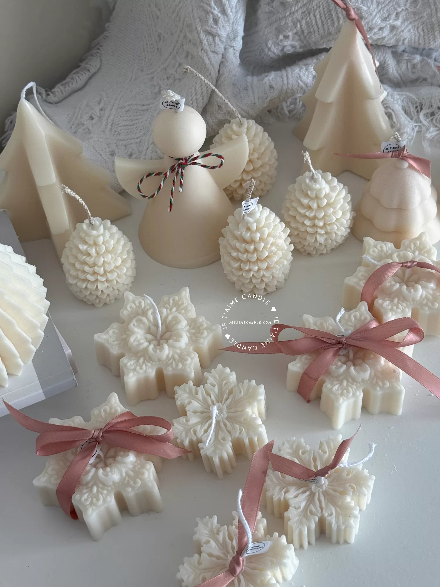 A collection of decorative candles in various shapes, including snowflakes, a Christmas tree, a nutcracker, a house, and a cylindrical candle. The snowflake candles are intricately designed in white, displayed on soft fabric.