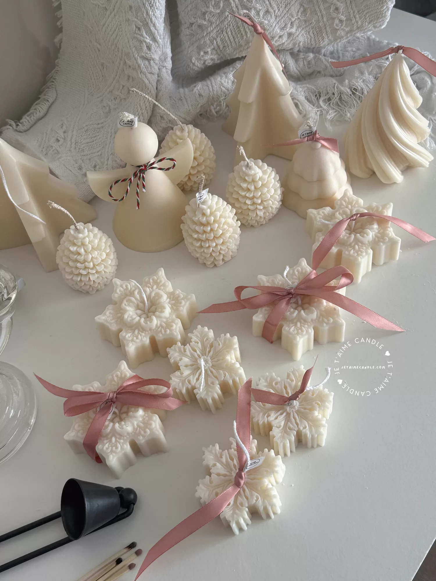 A collection of decorative candles in various shapes, including snowflakes, a Christmas tree, a nutcracker, a house, and a cylindrical candle. The snowflake candles are intricately designed in white, displayed on soft fabric.