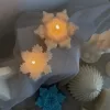 A collection of decorative candles in various shapes, including snowflakes, a Christmas tree, a nutcracker, a house, and a cylindrical candle. The snowflake candles are intricately designed in white and blue, displayed on soft fabric.