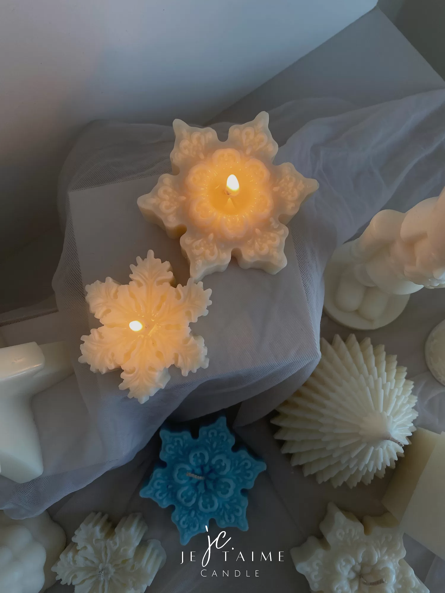 A collection of decorative candles in various shapes, including snowflakes, a Christmas tree, a nutcracker, a house, and a cylindrical candle. The snowflake candles are intricately designed in white and blue, displayed on soft fabric.