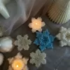 A collection of decorative candles in various shapes, including snowflakes, a Christmas tree, a nutcracker, a house, and a cylindrical candle. The snowflake candles are intricately designed in white and blue, displayed on soft fabric.