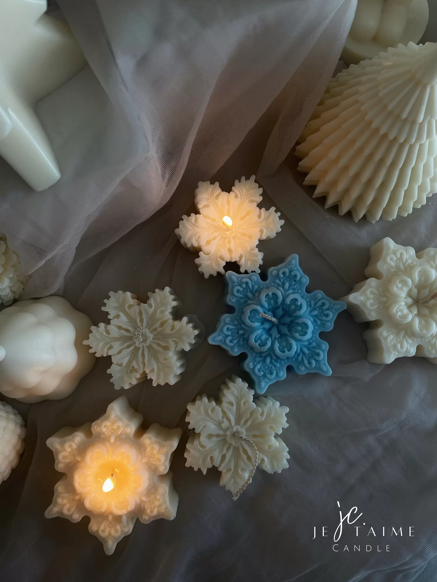 A collection of decorative candles in various shapes, including snowflakes, a Christmas tree, a nutcracker, a house, and a cylindrical candle. The snowflake candles are intricately designed in white and blue, displayed on soft fabric.