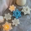 A collection of decorative candles in various shapes, including snowflakes, a Christmas tree, a nutcracker, a house, and a cylindrical candle. The snowflake candles are intricately designed in white and blue, displayed on soft fabric.