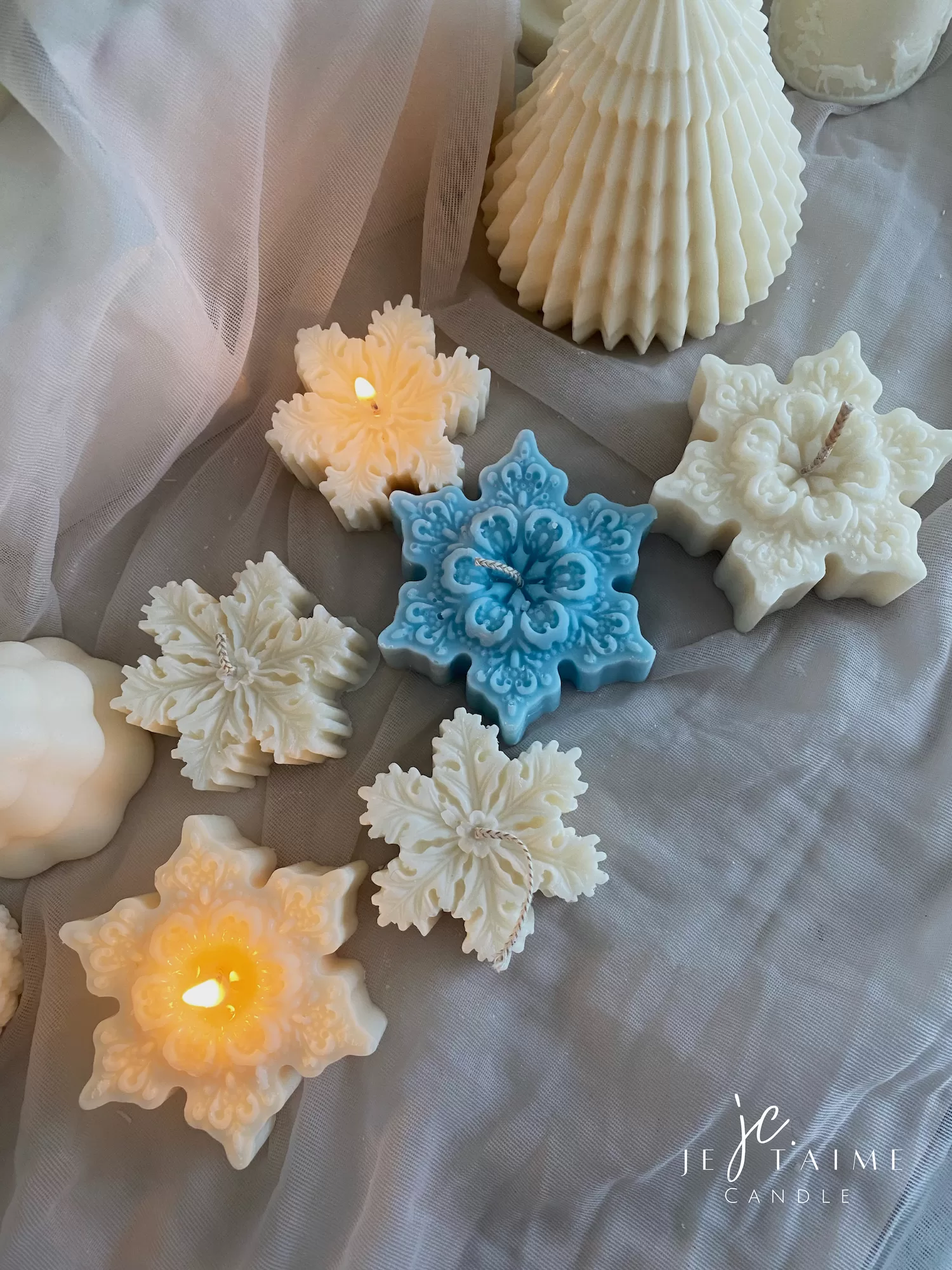A collection of decorative candles in various shapes, including snowflakes, a Christmas tree, a nutcracker, a house, and a cylindrical candle. The snowflake candles are intricately designed in white and blue, displayed on soft fabric.