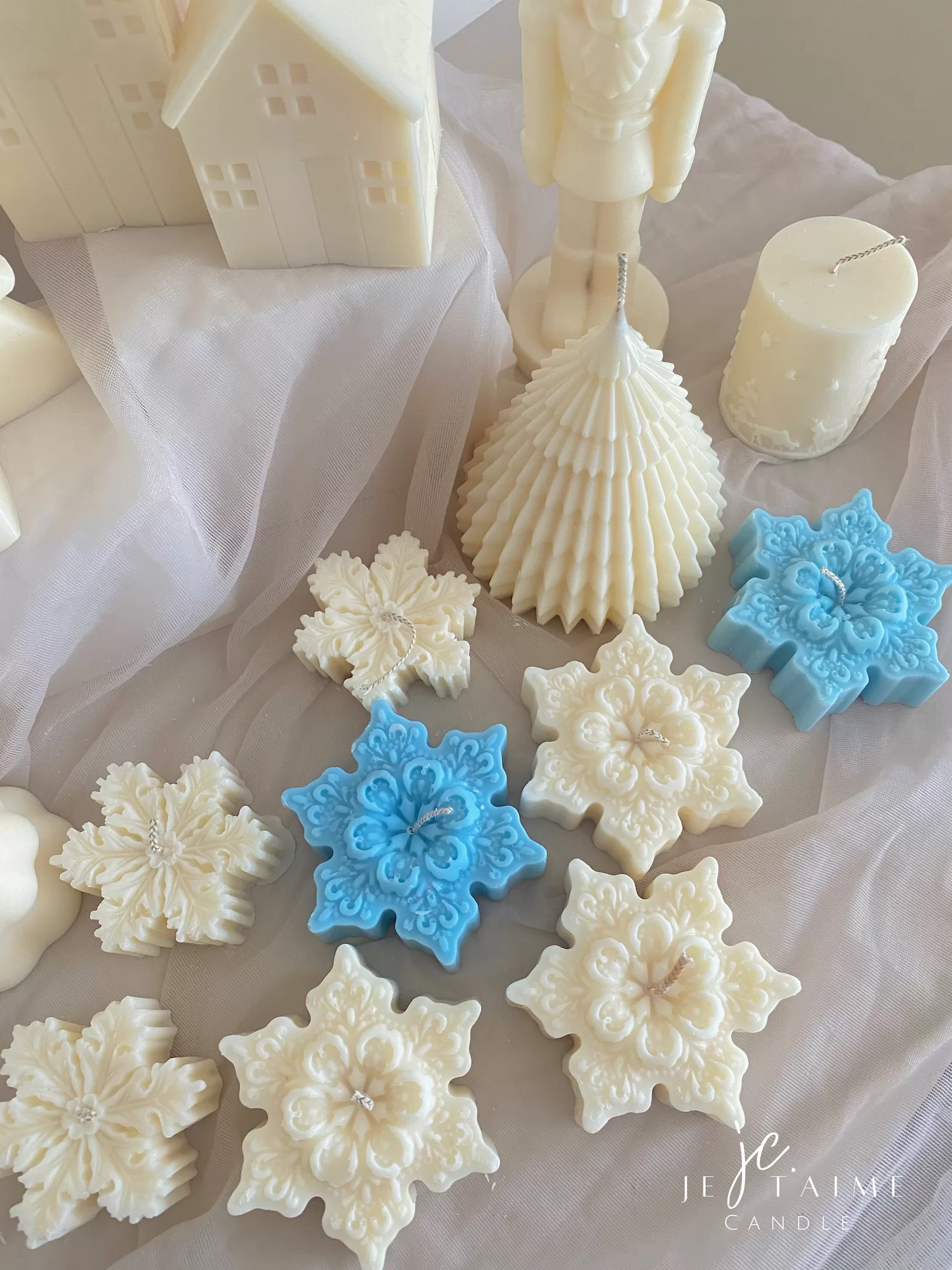 A collection of decorative candles in various shapes, including snowflakes, a Christmas tree, a nutcracker, a house, and a cylindrical candle. The snowflake candles are intricately designed in white and blue, displayed on soft fabric.