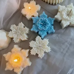 A collection of decorative candles in various shapes, including snowflakes, a Christmas tree, a nutcracker, a house, and a cylindrical candle. The snowflake candles are intricately designed in white and blue, displayed on soft fabric.