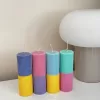 A row of tall ribbed pillar candles in pastel and bright two-tone colors, arranged on a white surface. The candles are displayed beside a green checkered lamp, a decorative mirror with an orange frame, and pampas grass in the background.