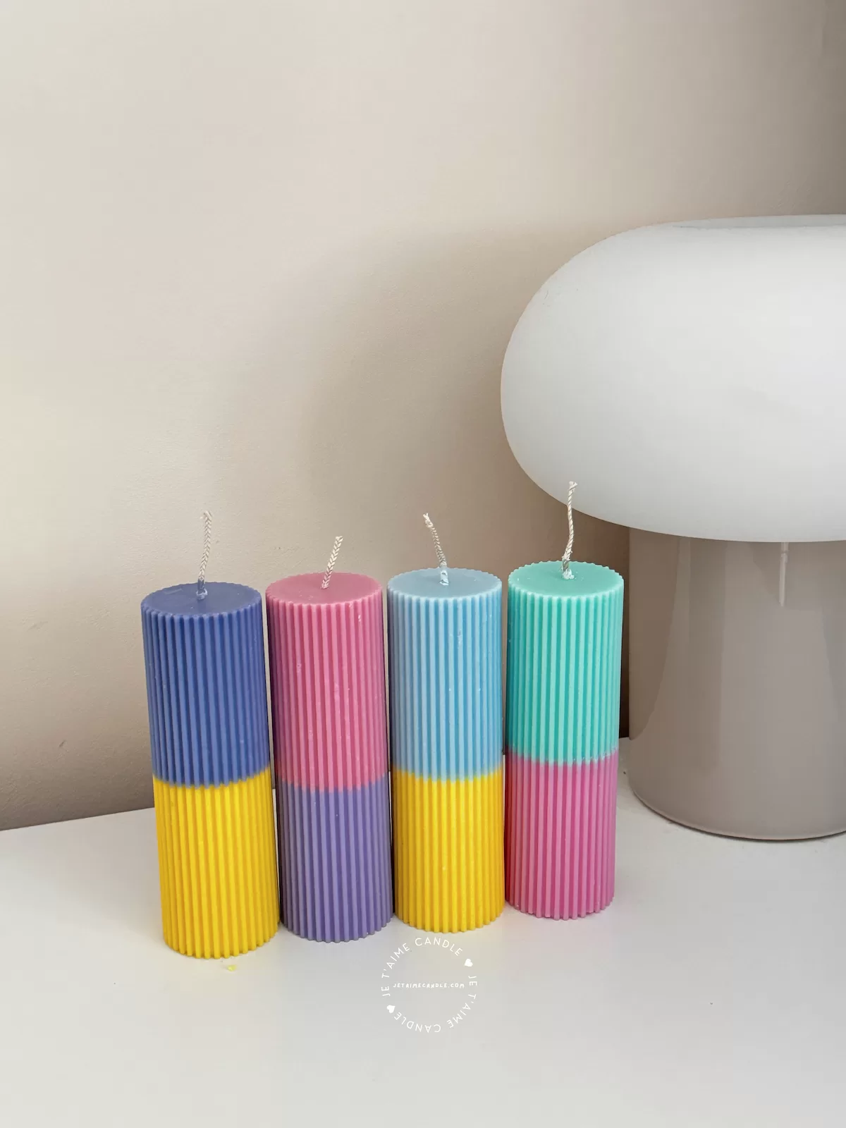 A row of tall ribbed pillar candles in pastel and bright two-tone colors, arranged on a white surface. The candles are displayed beside a green checkered lamp, a decorative mirror with an orange frame, and pampas grass in the background.