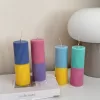 A row of tall ribbed pillar candles in pastel and bright two-tone colors, arranged on a white surface. The candles are displayed beside a green checkered lamp, a decorative mirror with an orange frame, and pampas grass in the background.