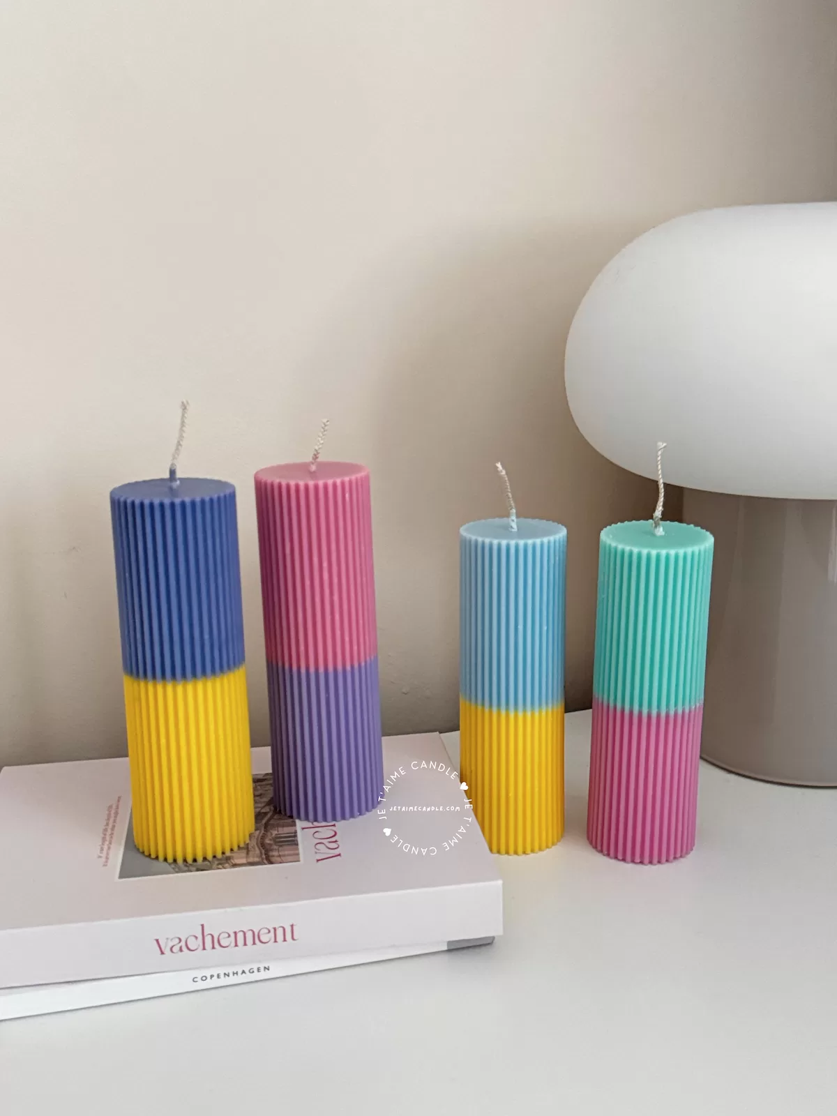 A row of tall ribbed pillar candles in pastel and bright two-tone colors, arranged on a white surface. The candles are displayed beside a green checkered lamp, a decorative mirror with an orange frame, and pampas grass in the background.