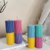 A row of tall ribbed pillar candles in pastel and bright two-tone colors, arranged on a white surface. The candles are displayed beside a green checkered lamp, a decorative mirror with an orange frame, and pampas grass in the background.