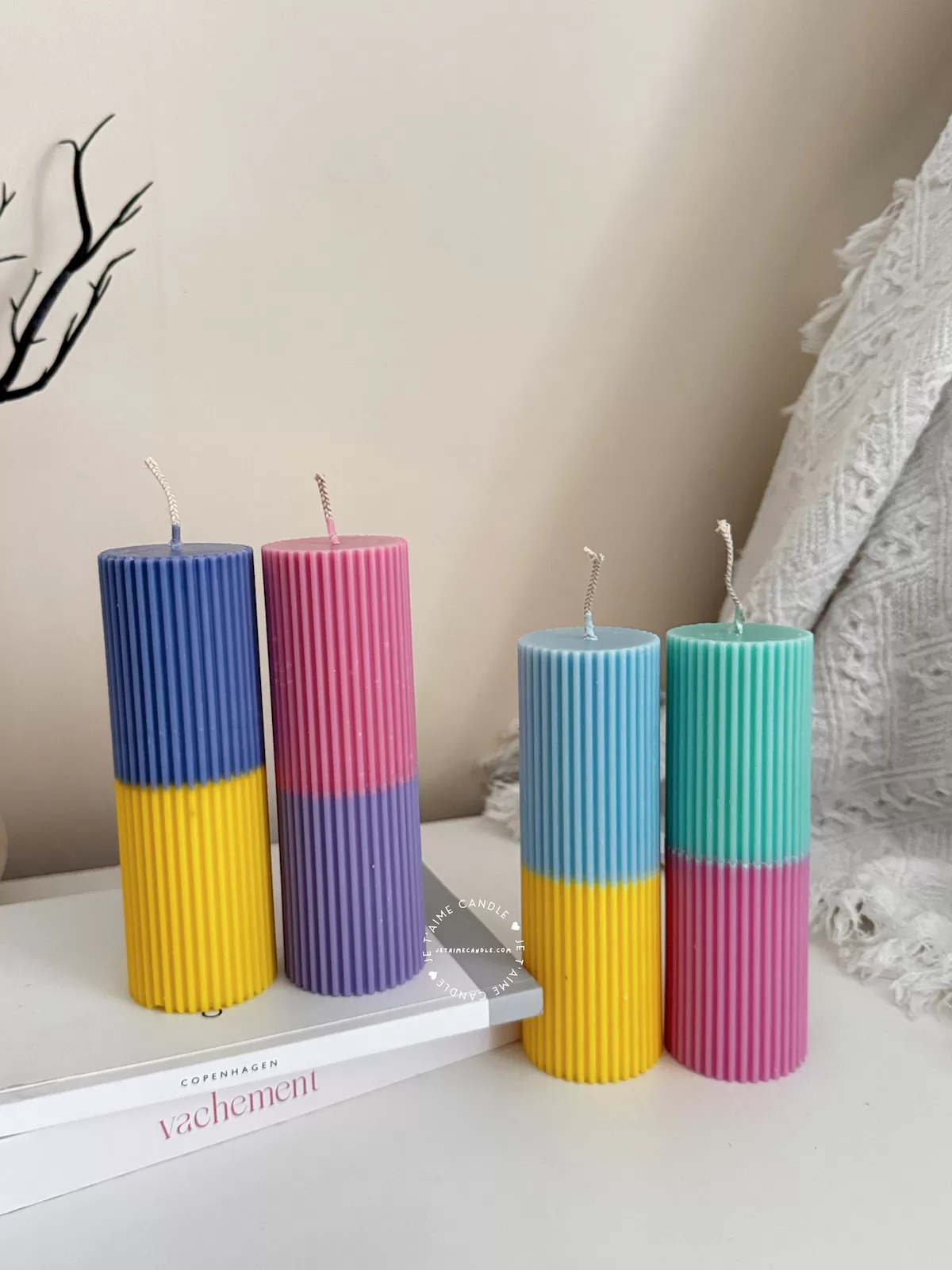 A row of tall ribbed pillar candles in pastel and bright two-tone colors, arranged on a white surface. The candles are displayed beside a green checkered lamp, a decorative mirror with an orange frame, and pampas grass in the background.