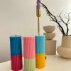 A row of tall ribbed pillar candles in pastel and bright two-tone colors, arranged on a white surface. The candles are displayed beside a green checkered lamp, a decorative mirror with an orange frame, and pampas grass in the background.