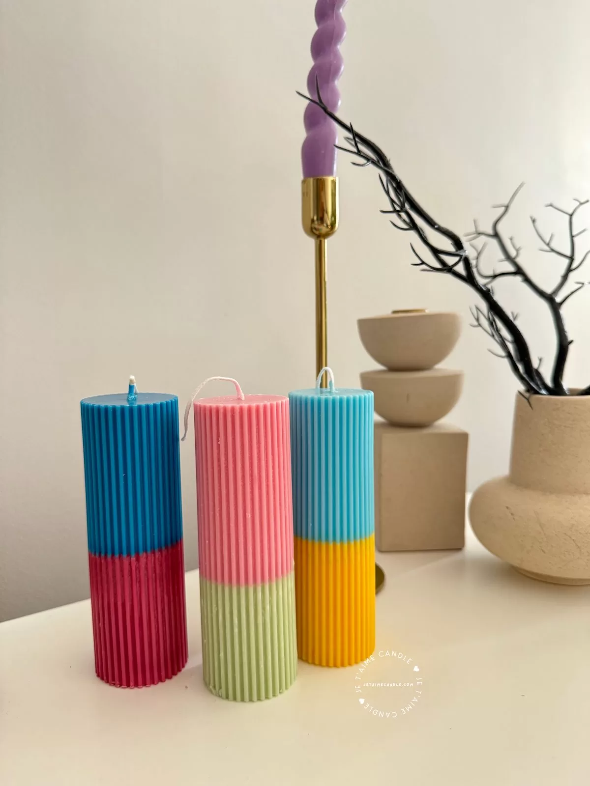 A row of tall ribbed pillar candles in pastel and bright two-tone colors, arranged on a white surface. The candles are displayed beside a green checkered lamp, a decorative mirror with an orange frame, and pampas grass in the background.
