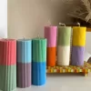 A row of tall ribbed pillar candles in pastel and bright two-tone colors, arranged on a white surface. The candles are displayed beside a green checkered lamp, a decorative mirror with an orange frame, and pampas grass in the background.