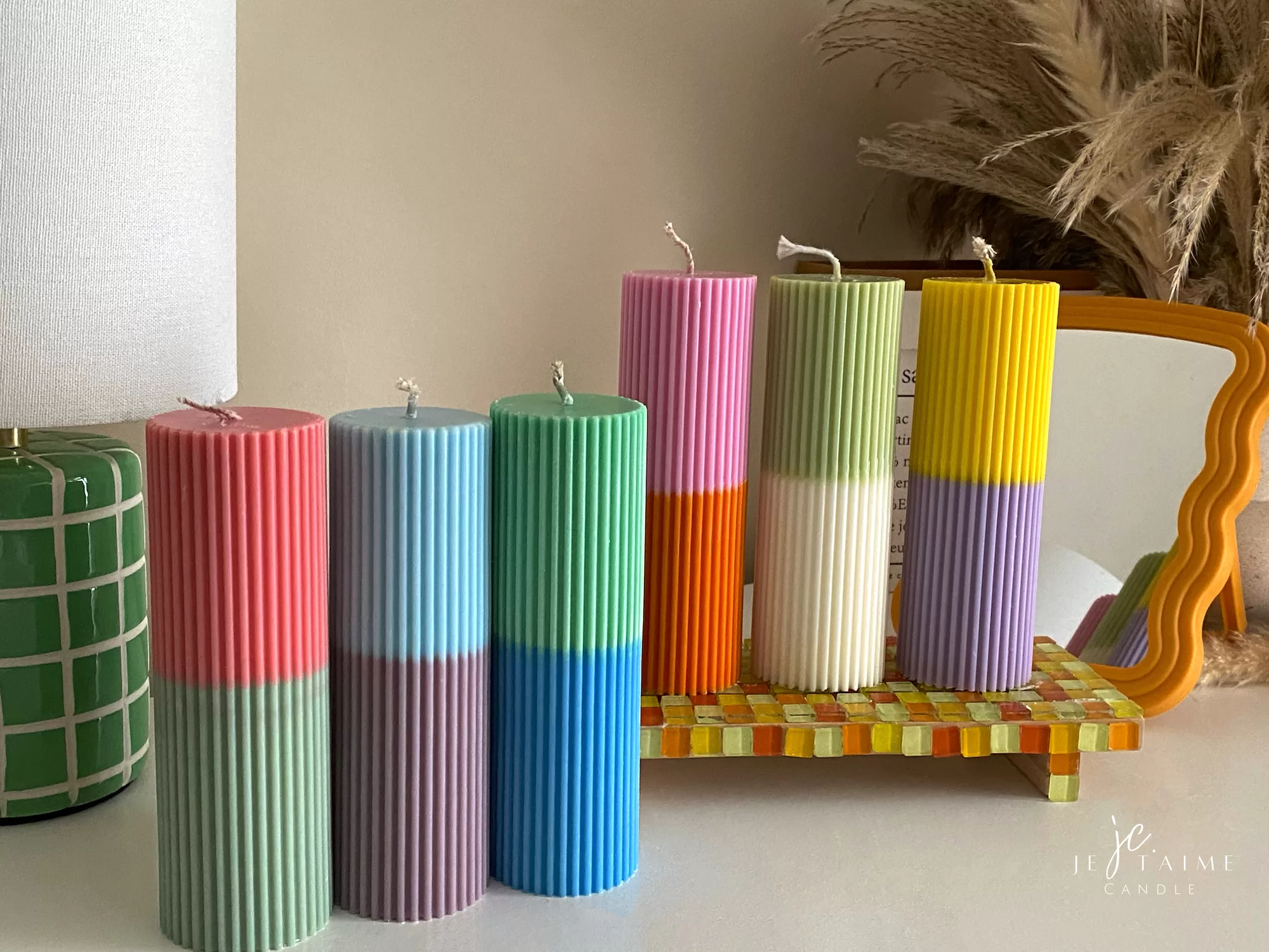 A row of tall ribbed pillar candles in pastel and bright two-tone colors, arranged on a white surface. The candles are displayed beside a green checkered lamp, a decorative mirror with an orange frame, and pampas grass in the background.