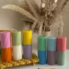 A row of tall ribbed pillar candles in pastel and bright two-tone colors, arranged on a white surface. The candles are displayed beside a green checkered lamp, a decorative mirror with an orange frame, and pampas grass in the background.